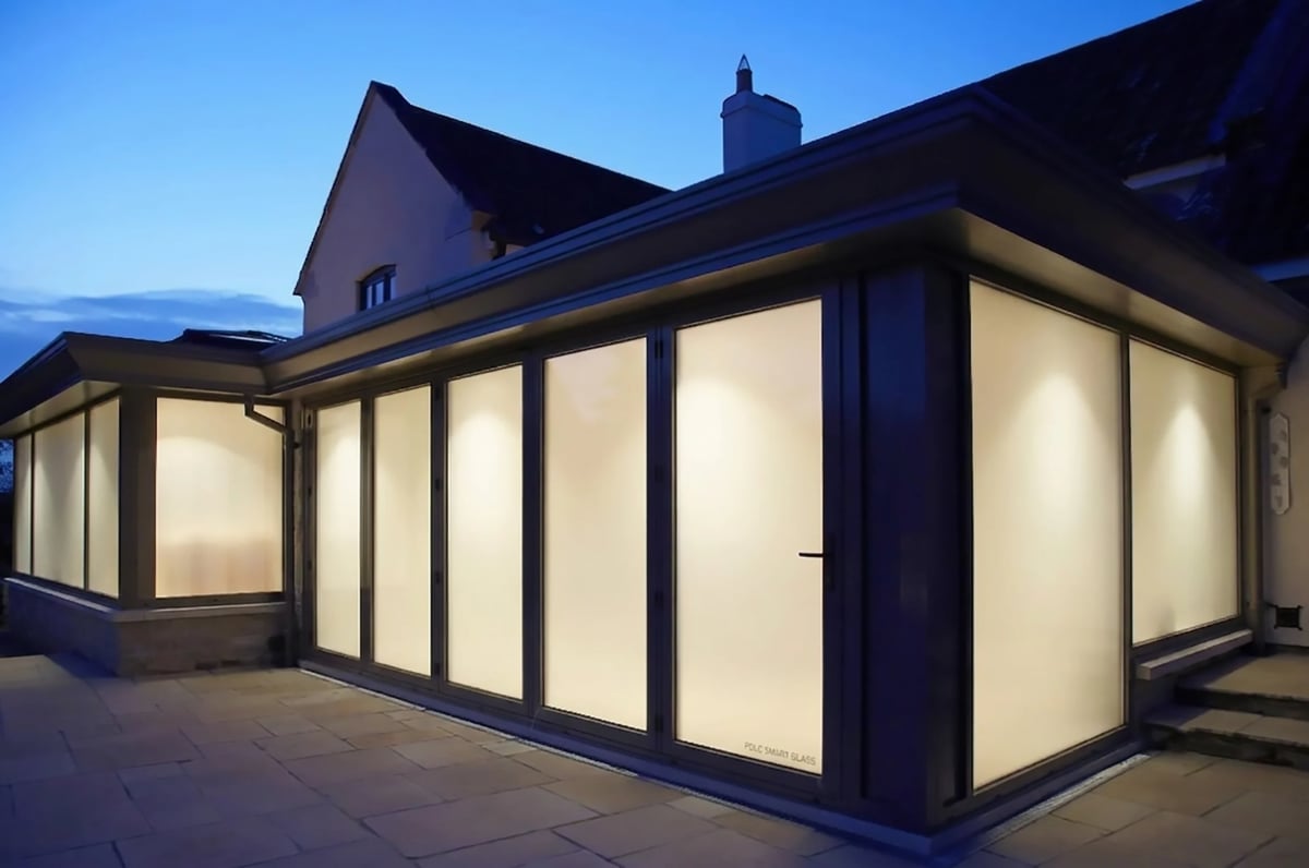 Modern glass-walled garden room extension lit from within at dusk, attached to a residential home with dark trim and roofline