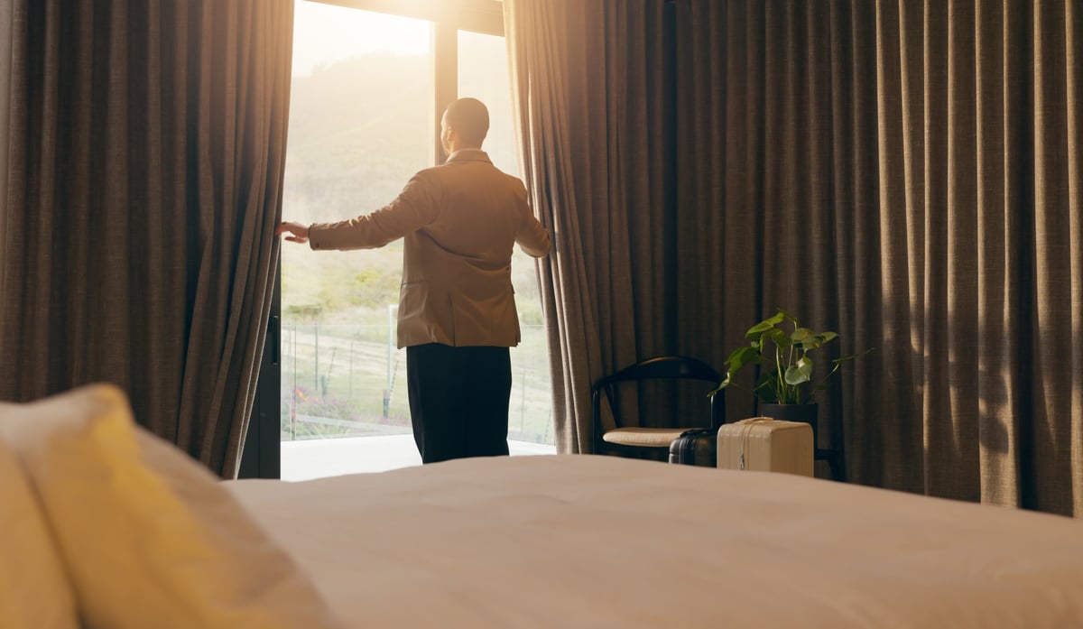 Person opening curtains in a bright bedroom with morning sunlight streaming through large windows