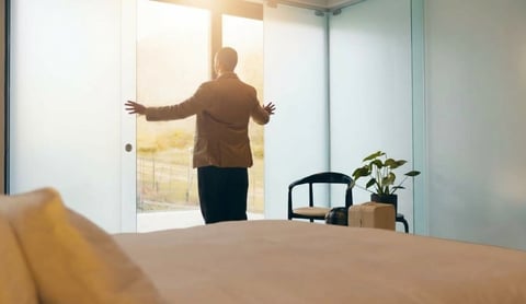 Man in brown jacket standing in modern office with arms outstretched, facing glass windows with sunlight