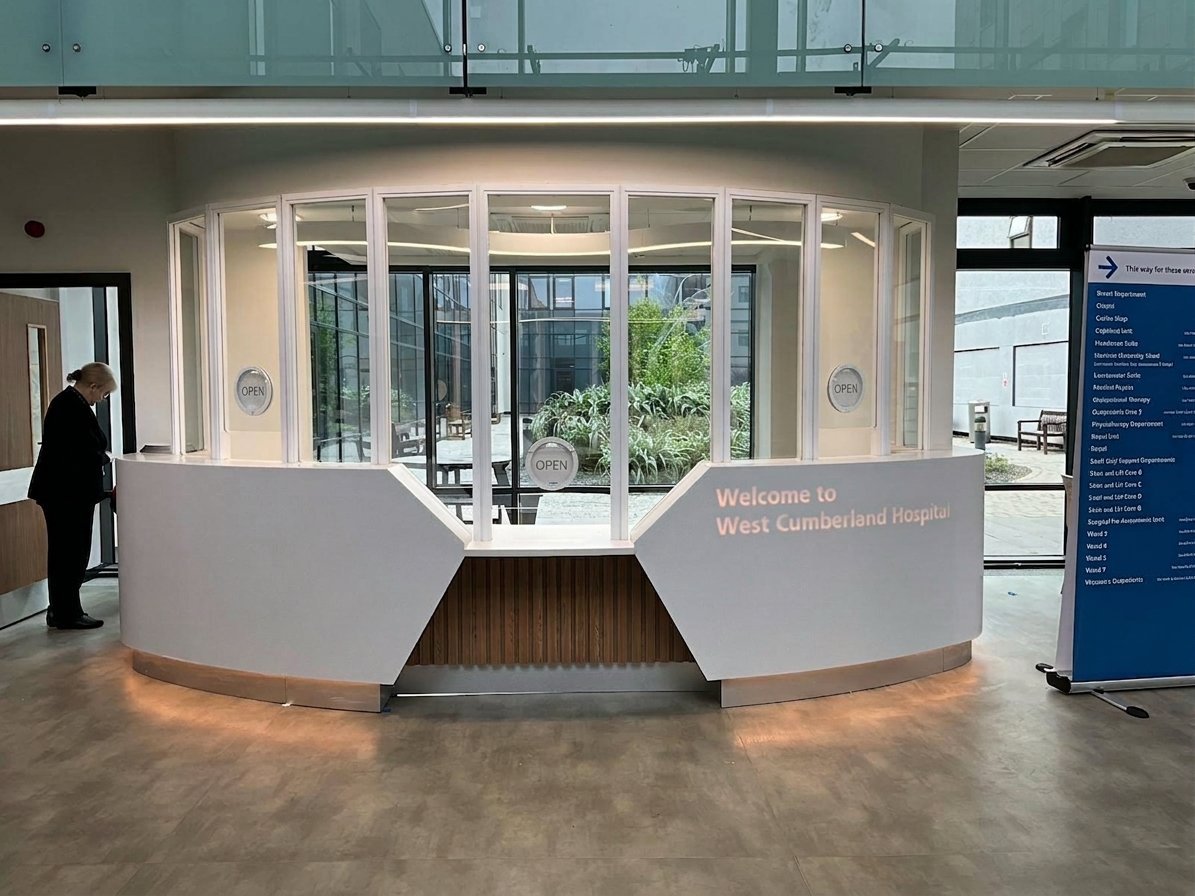 Modern curved white reception desk in a bright lobby with glass doors, indoor plants, and informational signage visible in the background