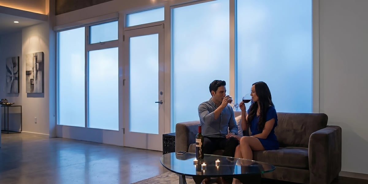 Two people sitting on a dark couch having a conversation in a modern living room with large windows and blue evening light